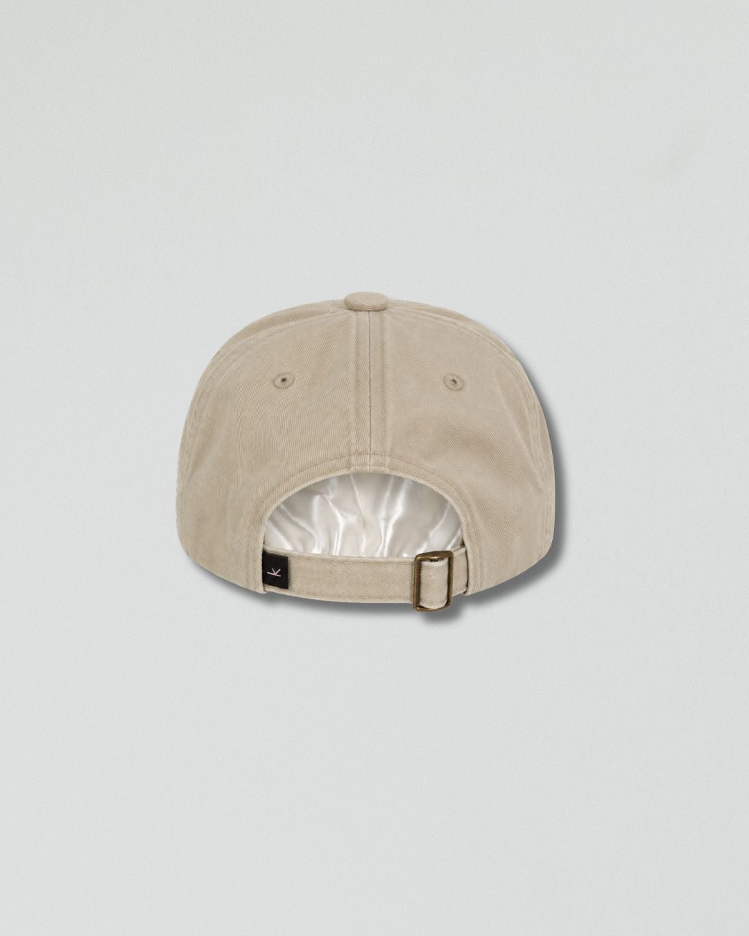 A light beige baseball cap shown from the back, featuring a fabric-covered metal buckle and a small black brand tag on the side, set against a solid light gray background.