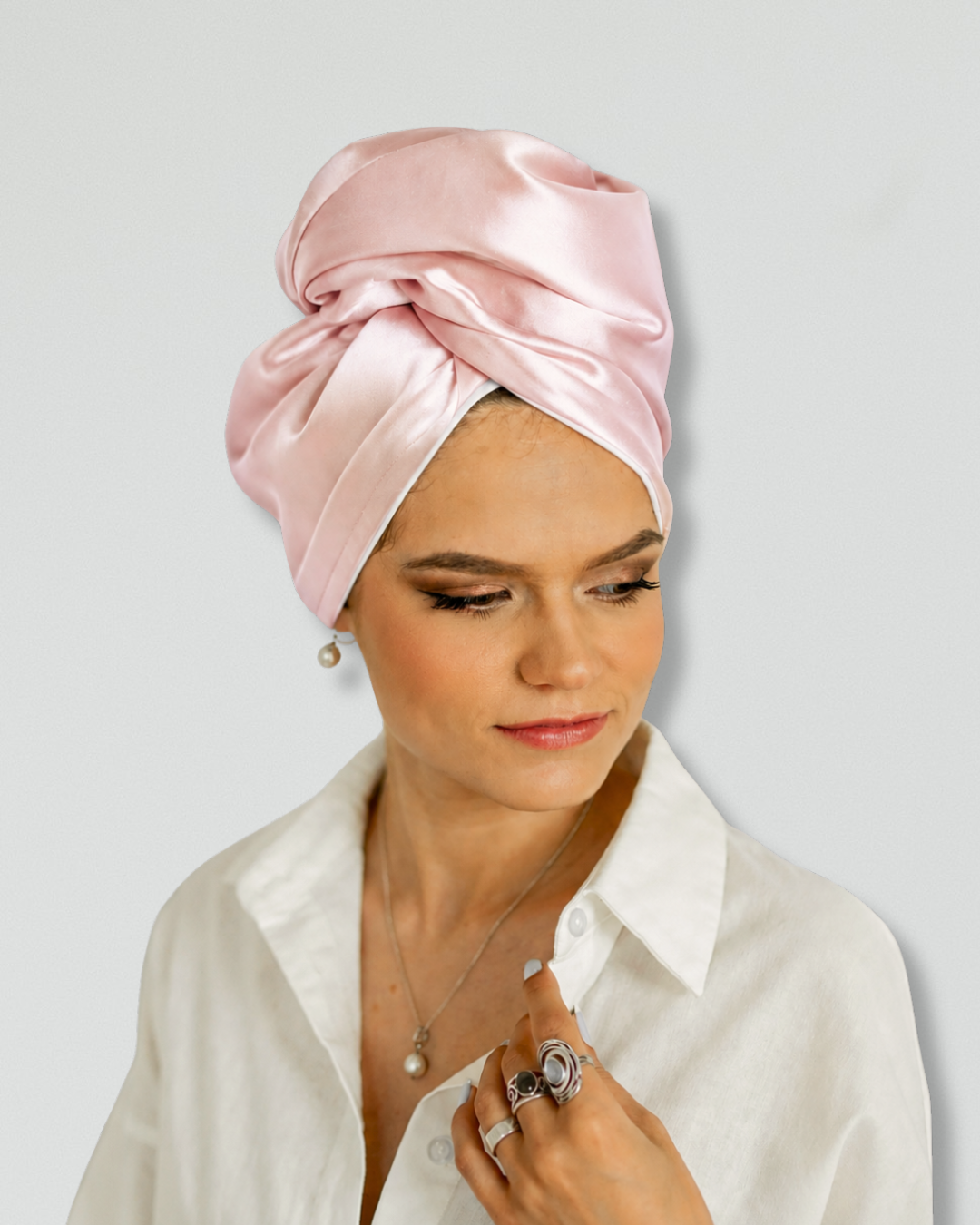 A woman wearing a light pink siilk turban headwrap and a white collared shirt, photographed against a plain light gray background.