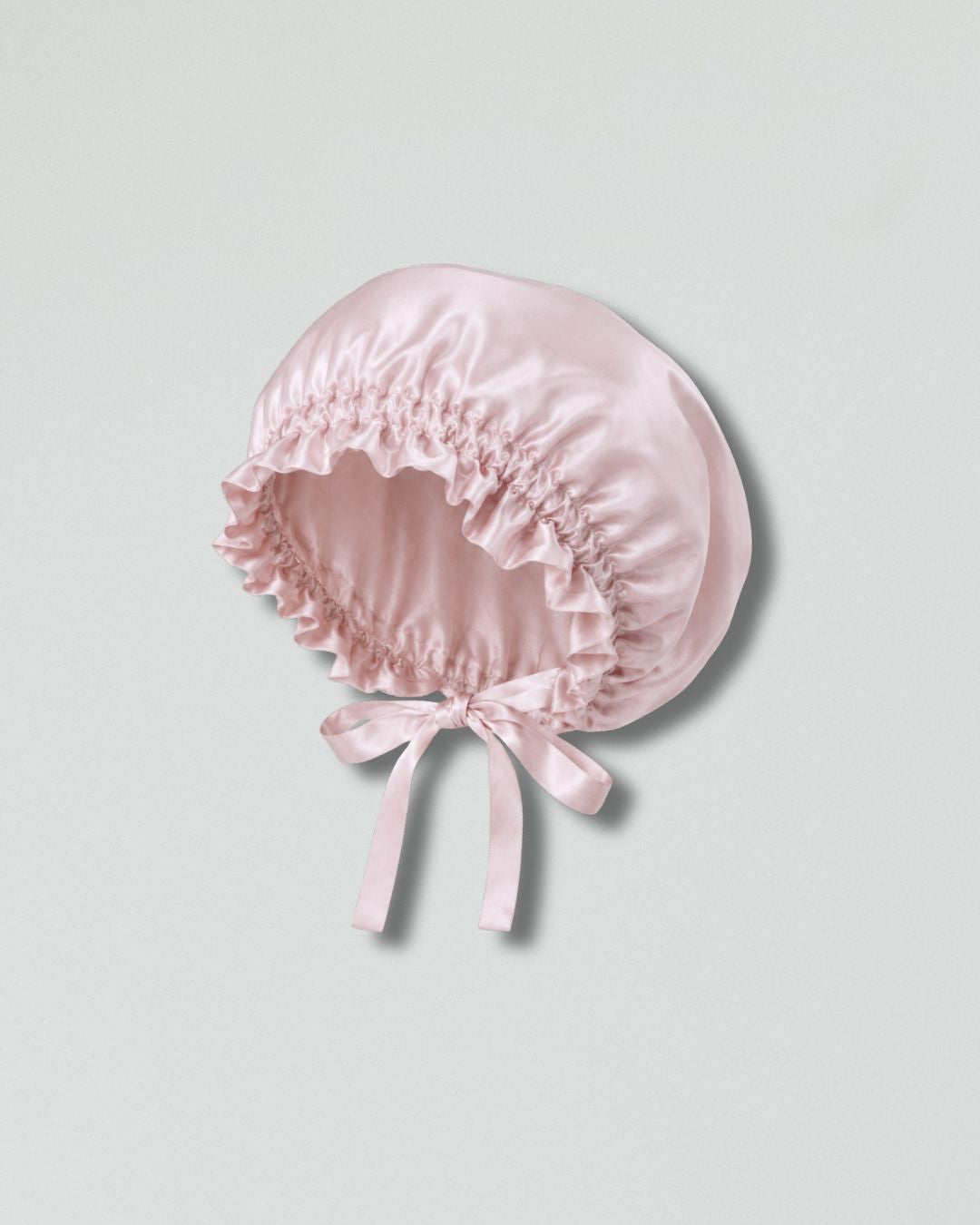 A light pink silk bonnet with a gathered ruffled brim and a matching bow at the front, displayed on a neutral light gray background.