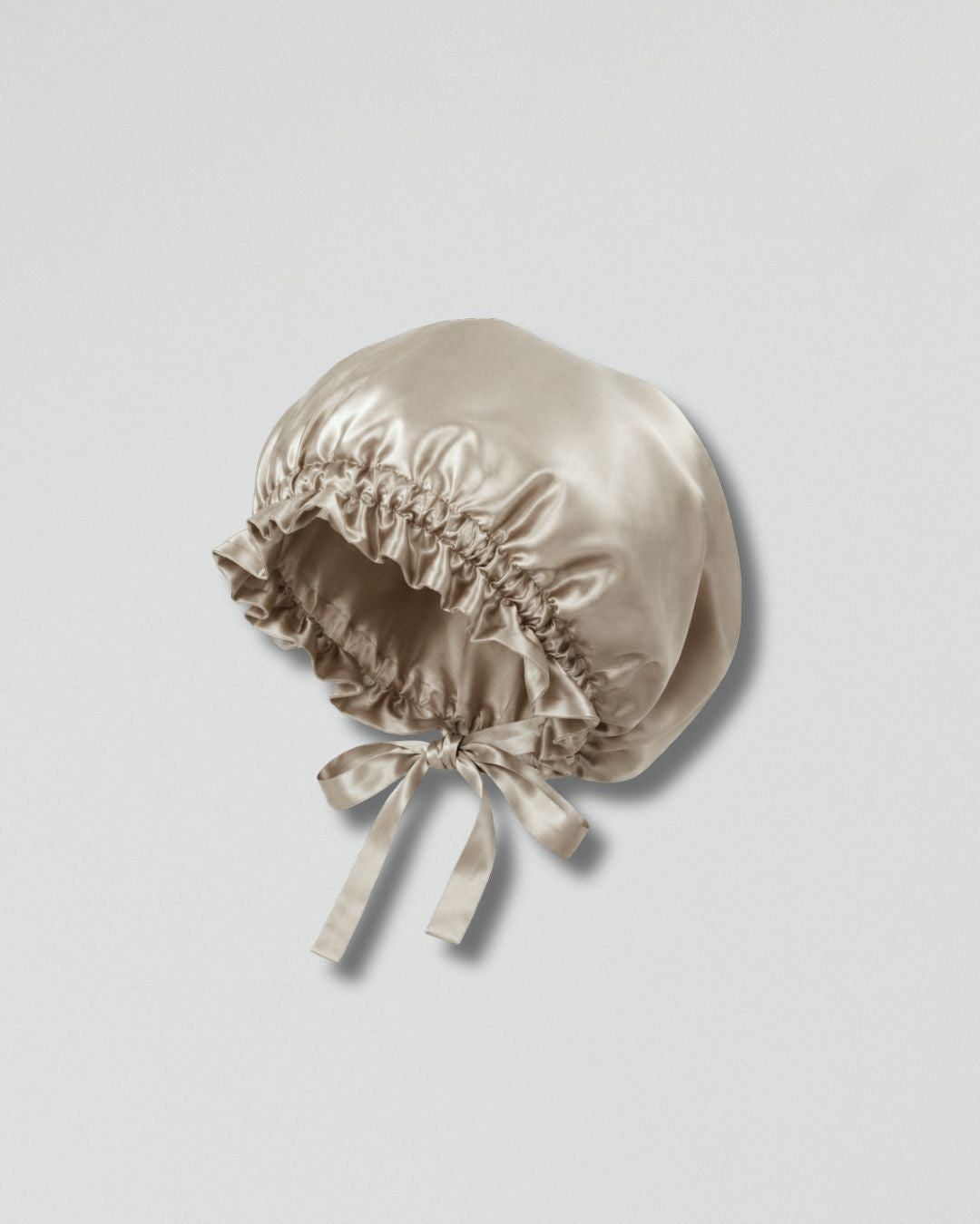 A champagne beige silk bonnet with a ruffled edge and a tied bow, shown on a neutral light gray background.