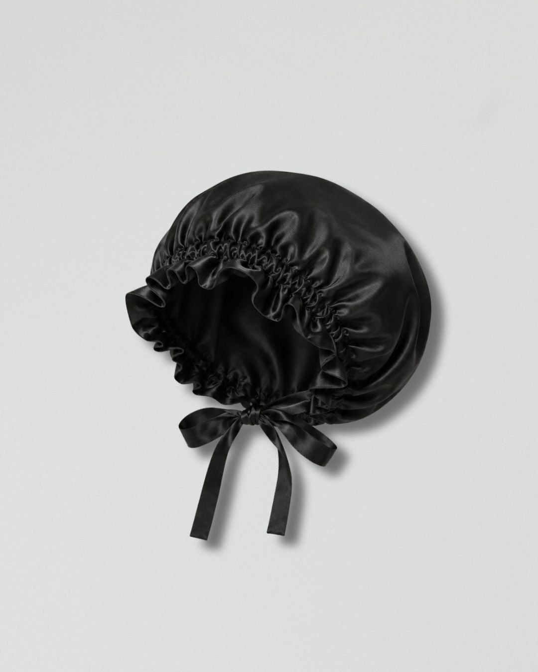 A black silk bonnet with a gathered ruffled edge and a matching satin bow, displayed on a light gray background.