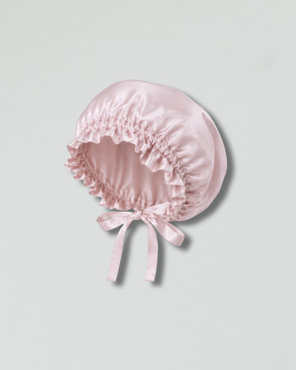 A light pink silk bonnet with a gathered ruffled brim and a matching bow at the front, displayed on a neutral light gray background.